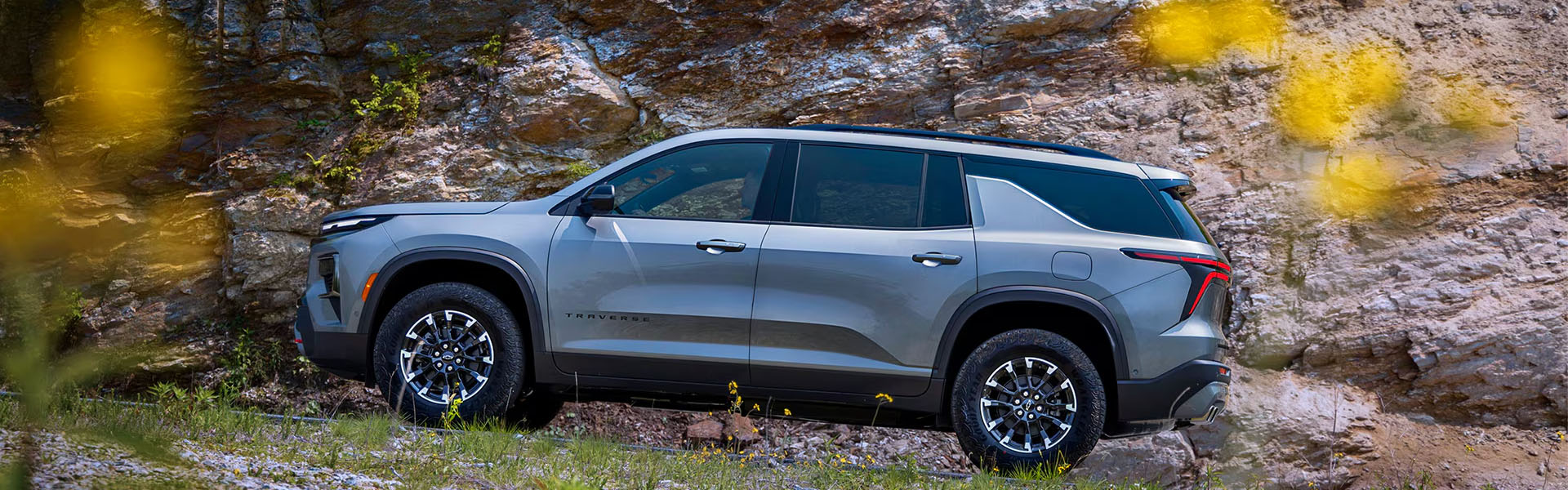 Profile view of a gray 2026 Chevrolet Traverse parked near rocky terrain with black wheels and natural surroundings.