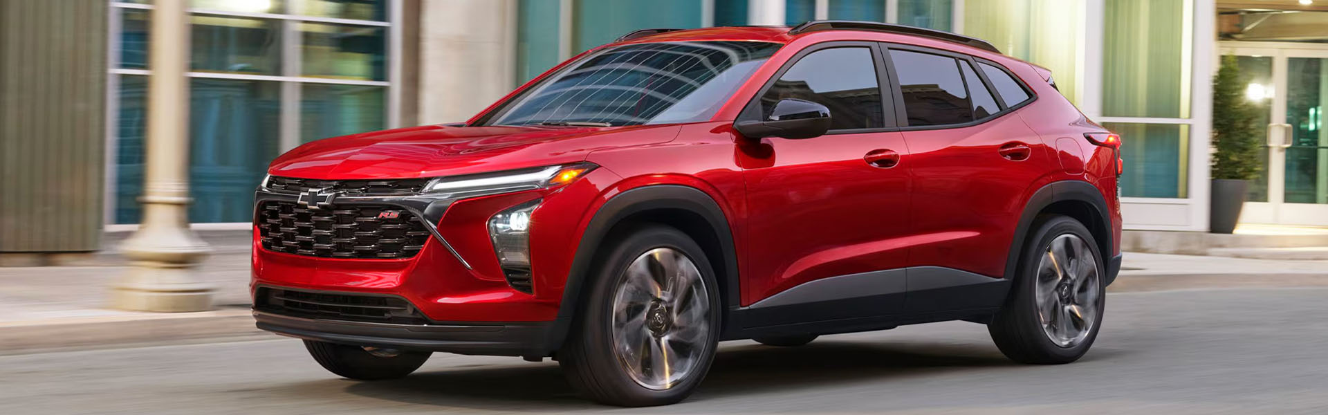 Red 2026 Chevrolet Trax driving through a modern city street with buildings in the background.