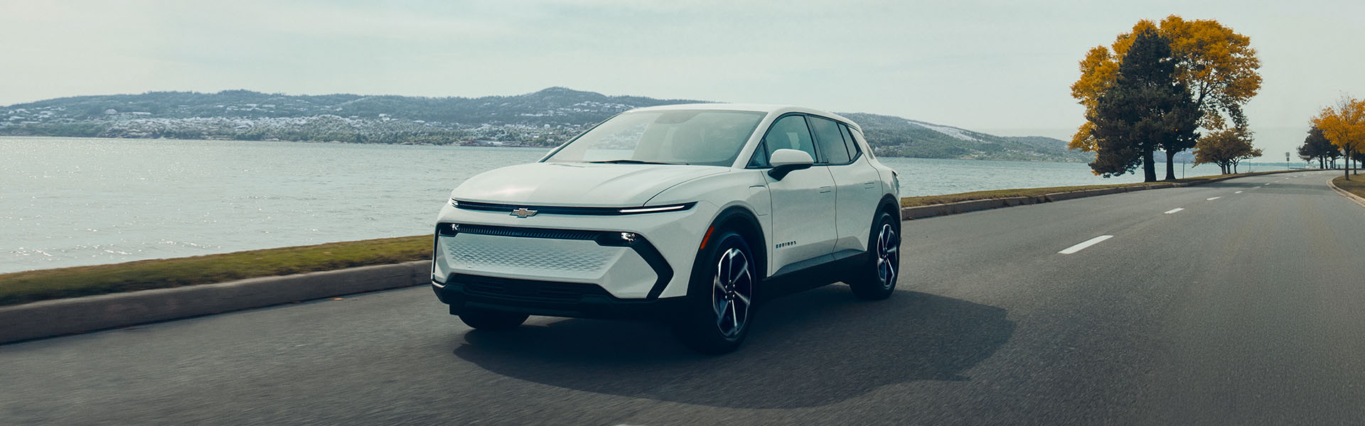 2026 Chevrolet Equinox EV driving along a lakeside road with green trees in the background.