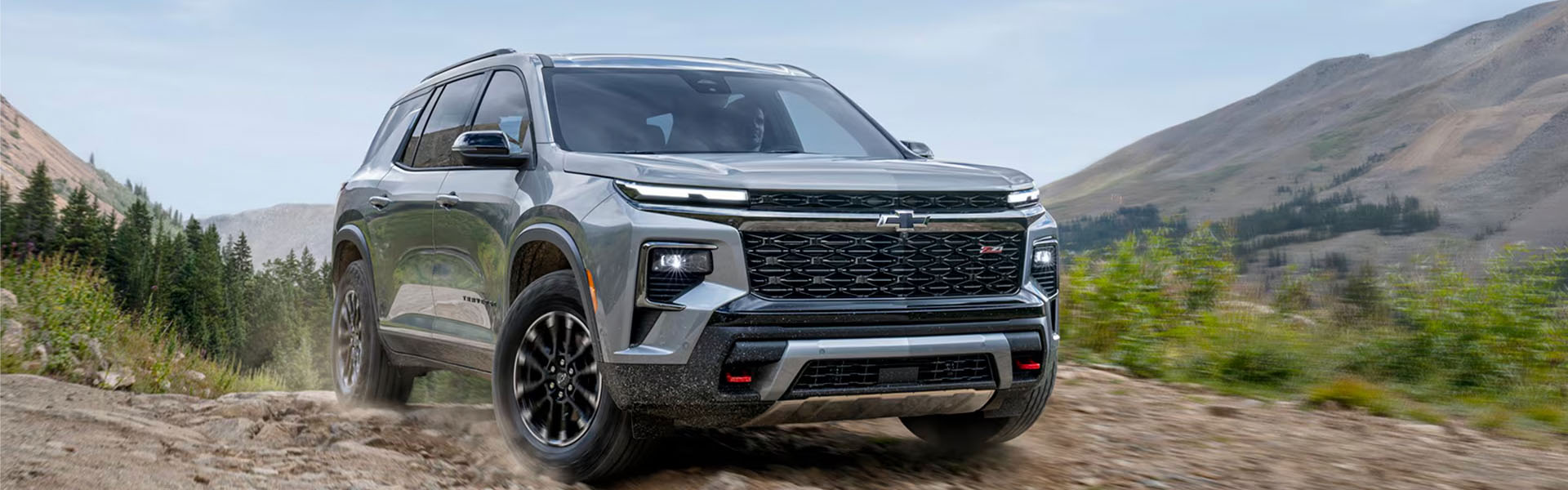 Silver 2026 Chevrolet Traverse driving off-road with mountains in the background.