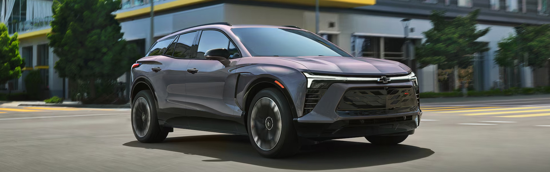 2026 Chevrolet Blazer EV in gray driving through a city street, showcasing its sleek aerodynamic design, distinctive LED lighting, bold front grille, and modern electric SUV styling.