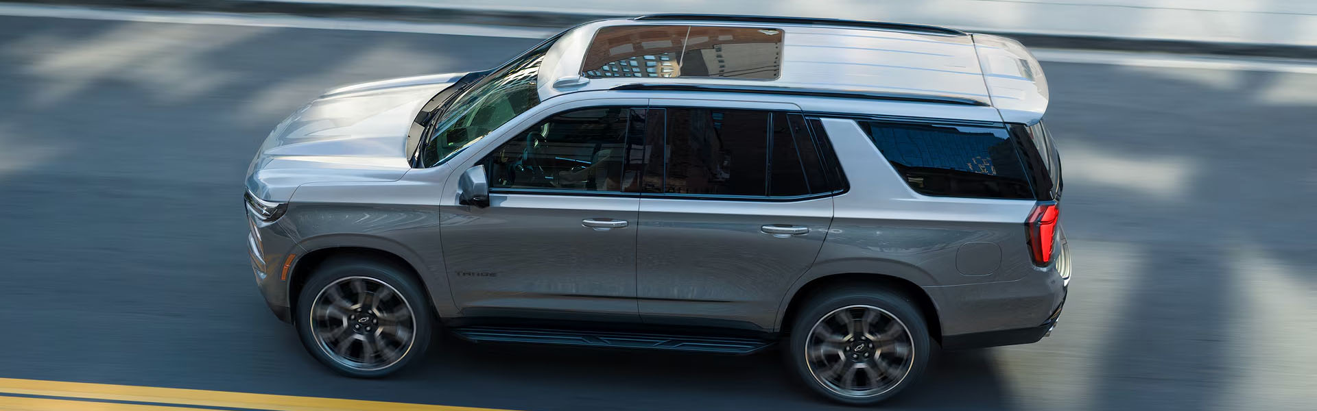 2026 Chevy Tahoe SUV driving on a city street, shown from an elevated side view with its spacious design, roof rails, and alloy wheels visible.